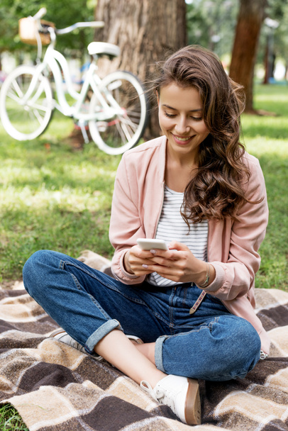 cheerful girl looking at smartphone while sitting on checkered blanket in park  - Foto, Bild