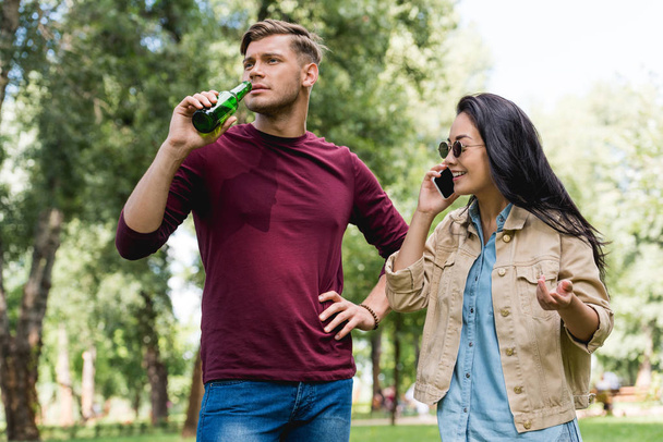 cheerful girl in sunglasses talking on smartphone near boyfriend drinking beer in park - Photo, Image