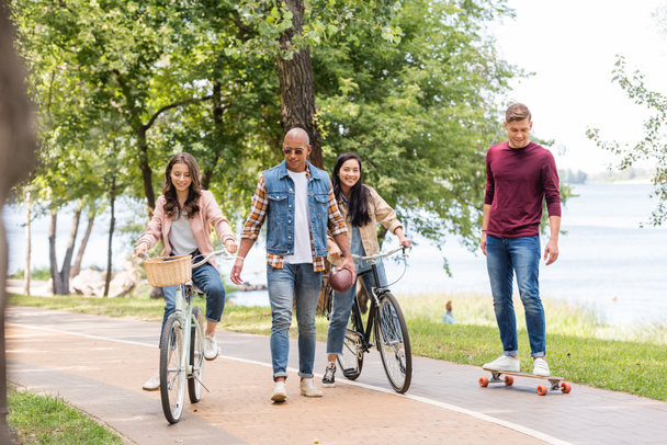 guapo afroamericano hombre caminando cerca de chicas guapas montando bicicletas y alegre amigo longboarding en parque
  - Foto, imagen