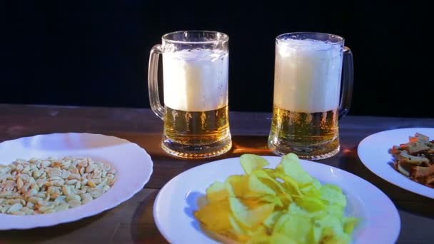 Dos tazas con cerveza espumosa sobre una mesa de madera en un bar sobre un fondo negro. Cerca de la placa con patatas fritas y nueces
 - Imágenes, Vídeo