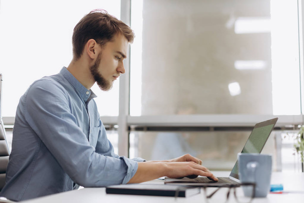 Volle Konzentration bei der Arbeit. schöner junger Mann mit Bart im Hemd arbeitet am Laptop, während er an seinem Arbeitsplatz sitzt - Foto, Bild
