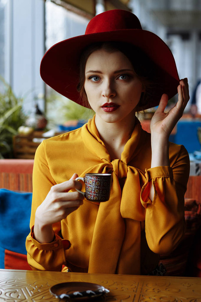 retrato de una chica en un café con una taza de café y un sombrero.retrato de una chica joven sensual con sombrero flojo y blusa con lazo. Hermosa mujer morena en el café sosteniendo la taza de café
 - Foto, imagen