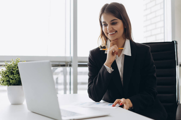 Kaunis liiketoiminnan nainen katselee kannettavaa tietokonetta ja hymyilee työskennellessään toimistossa. Keskittyminen työhön
. - Valokuva, kuva