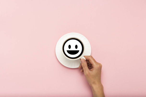 weibliche Hand, die eine weiße Tasse mit schwarzem Kaffee auf rosa Hintergrund hält. das Zeichen des Gesichts mit einem Lächeln. flache Lage, Draufsicht - Foto, Bild