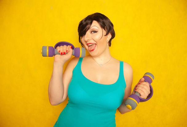 пухленькая радостная эмоциональная девушка с короткими волосами в синей футболке, занимающаяся спортом с гантелями, чтобы быть здоровой и похудеть на жёлтом фоне студии
 - Фото, изображение