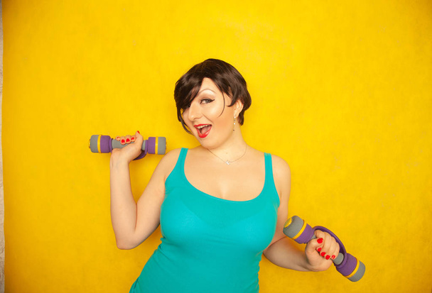 tombul neşeli duygusal kız mavi tişört kısa saçlı sağlıklı olmak ve kilo sarı katı studio arka plan üzerinde dumbbells ile spor yapıyor - Fotoğraf, Görsel