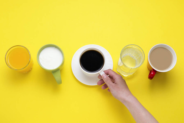 Female hand Holds Cup with Black coffee from a set of Drinks Orange juice, Coffee with Milk, Just Water, Yogurt on a yellow background. Flat lay, top view - Photo, Image