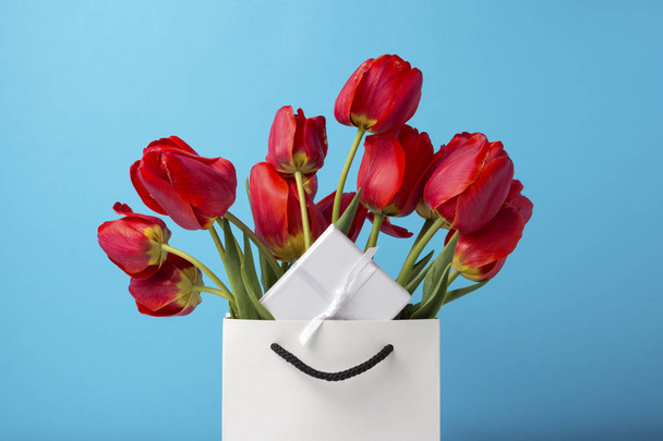 青色の背景に白いギフト袋に鮮やかな赤のチューリップの花束。お祝い、サプライズ、ギフトの概念 - 写真・画像