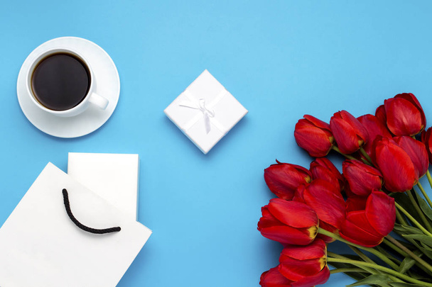 Bolsa de regalo blanca, una pequeña caja de regalo blanca, una taza blanca con café negro y un ramo de tulipanes rojos sobre un fondo azul. Concepto Ofrece un compromiso o matrimonio, compras
 - Foto, Imagen