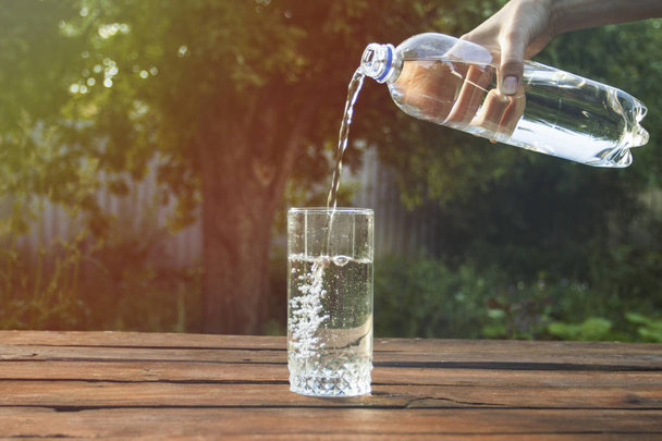 Naiskäsi kaataa vettä muovipullosta lasiin puupöydällä kevätpuutarhassa
 - Valokuva, kuva
