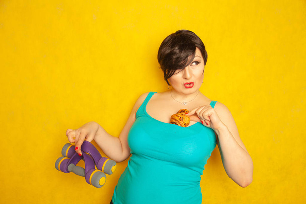 plump joyful emotional girl with short hair in a blue t-shirt tries to play sports with dumbbells to be healthy and lose weight, but can not refuse a croissant and harmful food - 写真・画像