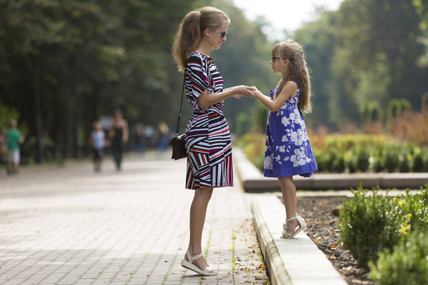 Young blond woman and small child girl in fashionable dresses holding hands on sunny park alley. - Photo, Image
