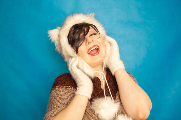 γοητευτικό κορίτσι Καυκάσιος σε ένα γούνινο καπέλο λευκό χειμώνα με γάτα αυτιά χαμόγελα και απολαμβάνει τη ζωή σε μπλε συμπαγές παρασκήνιο στο στούντιο μόνο - Φωτογραφία, εικόνα
