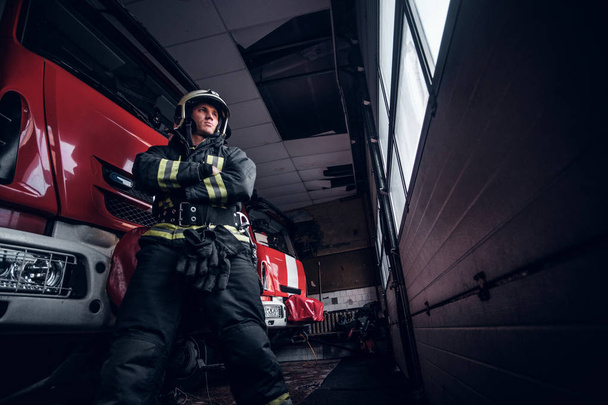 Χαμηλή γωνία φωτογραφία. Αυτοπεποίθηση πυροσβέστης που φοράει προστατευτική στολή στέκεται δίπλα σε ένα πυροσβεστικό όχημα σε ένα γκαράζ του μια πυροσβεστική - Φωτογραφία, εικόνα