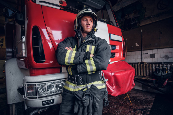 Jonge brandweerman dragen van beschermende uniforme staande naast een brandweerwagen in een garage van een brandweer - Foto, afbeelding