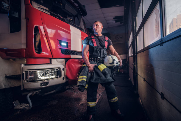 Gespierde brandweerman houden een beschermende helm in een garage van een brandweer - Foto, afbeelding