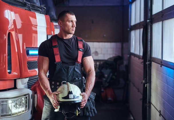 М'язистий пожежник тримає захисний шолом в гаражі пожежного відділення
 - Фото, зображення