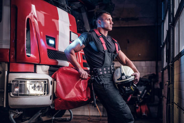 Мускулистый пожарный с защитным шлемом в гараже пожарной охраны
 - Фото, изображение