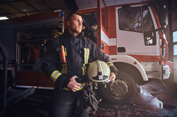 Bombero vistiendo uniforme mirando afuera mientras estaba parado cerca de un camión de bomberos en un garaje de un departamento de bomberos
 - Foto, imagen