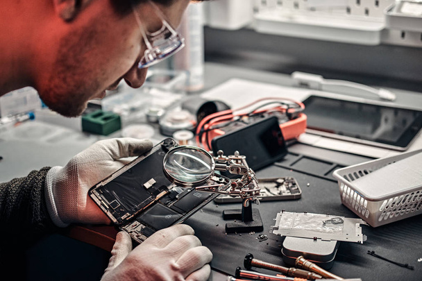 Technician uses a magnifying glass to carefully inspect the internal parts of the smartphone in a modern repair shop - Photo, Image