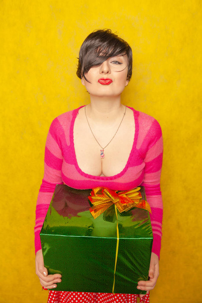 cute girl in pink striped sweater with big green gift with bow on yellow Studio background - Фото, изображение