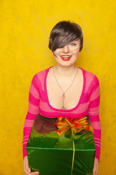 милая девушка в розовой полосатой свитер с большим зеленым подарком с луком на желтом фоне студии
 - Фото, изображение
