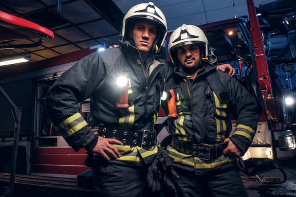 Двоє пожежників обіймаються і дивляться на камеру, що стоїть біля пожежної машини вночі на пожежному складі
 - Фото, зображення