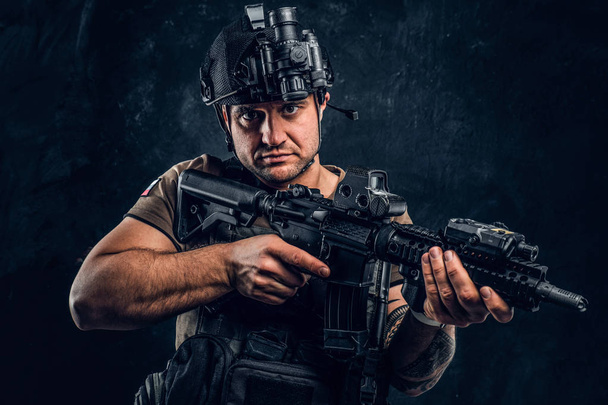 brutaler Soldat im T-Shirt mit Tätowierung auf der Hand, der Körperpanzer trägt und Helm mit Nachtsichtgerät, der mit einem Sturmgewehr posiert. - Foto, Bild