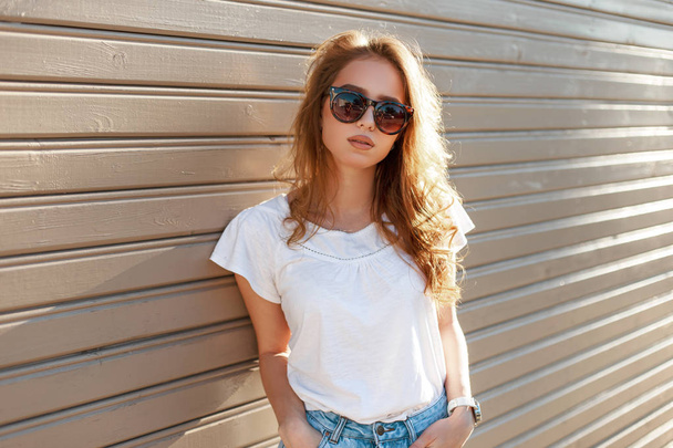 Young modern hipster woman in stylish sunglasses in a summer fashion white t-shirt in trendy jeans is standing near a wooden vintage wall on a sunny spring day. Cute girl resting outdoors. - Photo, Image
