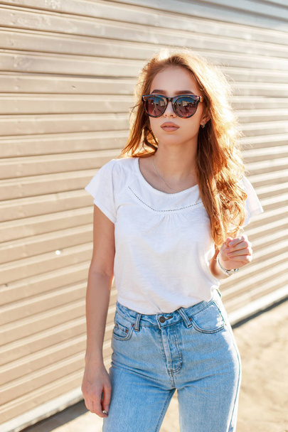 Stylish modern young hipster woman in fashionable sunglasses in white t-shirt in trendy jeans stands near a wooden vintage wall on a sunny warm day. Attractive American girl travels around the city. - Foto, Imagem