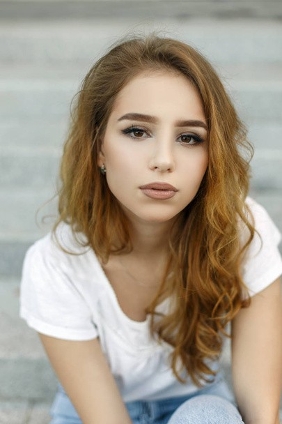 Portrait of pretty young stylish woman in fashionable white t-shirt in jeans with stylish hairstyle with beautiful eyes outdoors. Attractive girl resting on a warm summer day. - Valokuva, kuva