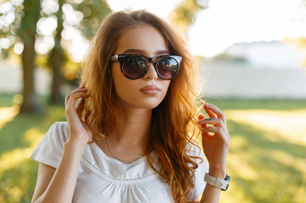Portrait d'une belle jolie jeune rousse hipster femme en lunettes de soleil tendance dans un t-shirt blanc à l'extérieur. Attrayant fille modèle promenades dans le parc par une chaude journée d'été et profite du soleil
. - Photo, image