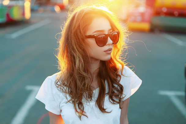 Portrait d'une jolie jolie jeune femme rousse hipster aux lèvres sexy avec des lunettes de soleil tendance en t-shirt blanc sur fond de coucher de soleil d'été orange vif. attrayant fille modèle bénéficie du soleil
. - Photo, image