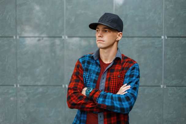Handsome stylish young man in a stylish T-shirt in a trendy blue and red plaid shirt in a black trendy basketball cap stands near a gray vintage wall. Cool guy. American style men's clothing. - Foto, Imagen