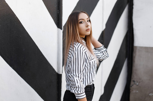 Pretty fashionable young woman in a vintage striped shirt with pierced nose in a velvet necklace in stylish black jeans posing near a black and white wall in stripes. Beautiful sexy girl model. - Photo, Image