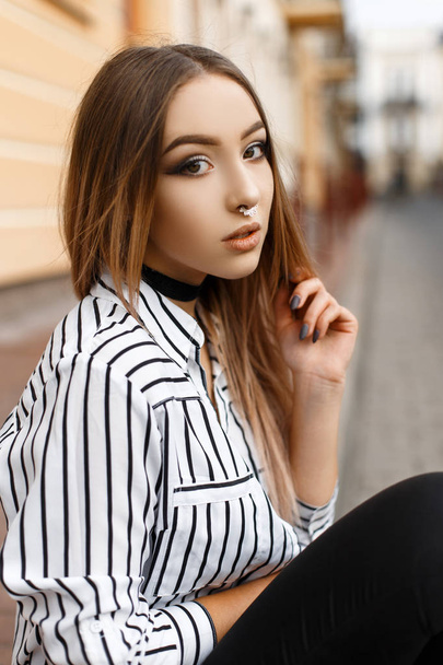Bela jovem atraente em um colar de veludo com um brinco no nariz em uma blusa listrada elegante em jeans preto perto de um edifício vintage. Menina adolescente moderna na moda ao ar livre
. - Foto, Imagem