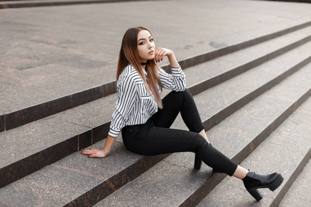 Довольно привлекательная молодая женщина в винтажной полосатой блузке в стильных джинсах в модных кожаных туфлях сидит на лестнице в городе в теплый весенний день. Стильная девушка-подросток расслабляется
. - Фото, изображение