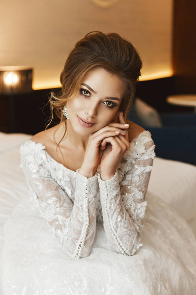 Portrait of beautiful and sensual brunette model girl with bright makeup and stylish hairstyle, in fashionable lace dress sits on a bed and posing at a luxury interior - Foto, Bild