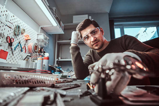 Technicien électronique en lunettes, appuyé sur un bureau dans un atelier de réparation, regardant une caméra avec un regard réfléchi
 - Photo, image