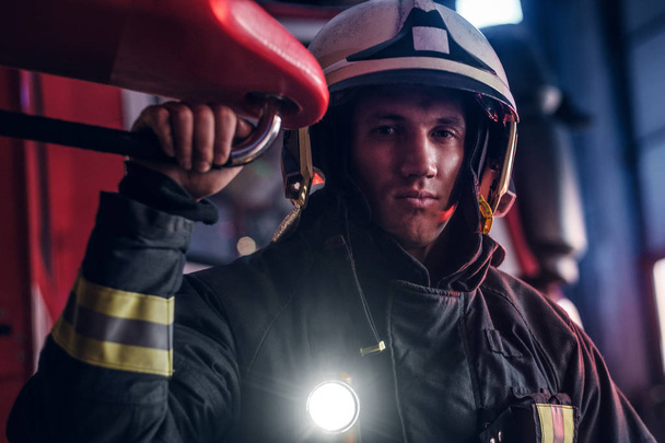 Retrato de um belo bombeiro vestindo um uniforme protetor com lanterna incluído em pé em uma garagem do quartel de bombeiros
 - Foto, Imagem