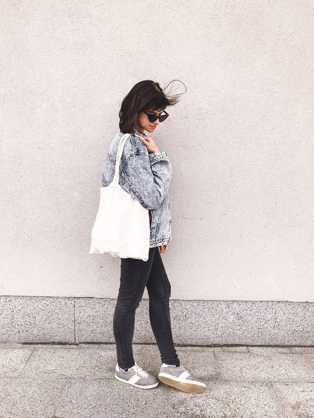 Elegante chica hipster en traje de mezclilla y bolso de mano posando en la ciudad
 - Foto, Imagen