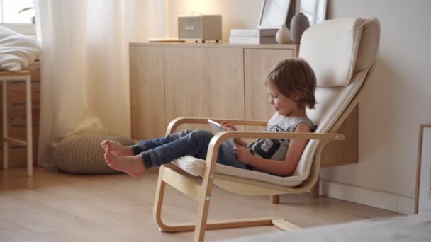 Cute 5 year caucasian boy sit in armchar and use tablet at light room - Felvétel, videó