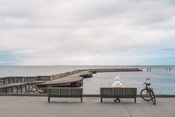 A young Caucasian woman sits with her back on a wooden bench overlooking the Baltic Sea on the seafront in Copenhagen Denmark in winter in cloudy weather. Girl walking gonoskoy bike parked nearby - Photo, image