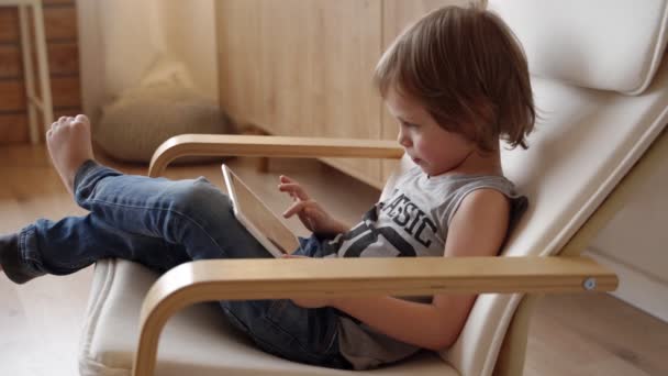Cute 5 year caucasian boy sit in armchar and use tablet at light room - Imágenes, Vídeo