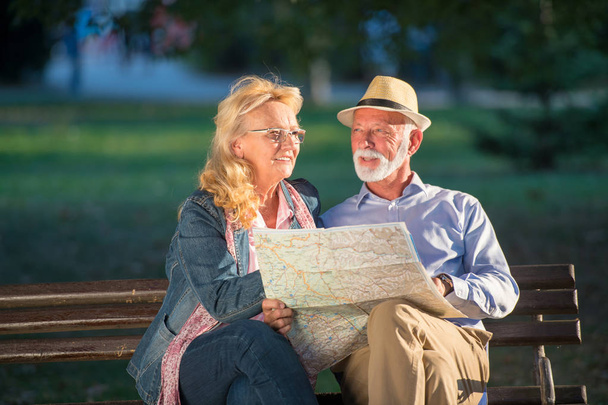 Portrait of happy man and woman reading map while sitting on a park bench. Senior couple on vacation using city map. - Photo, image