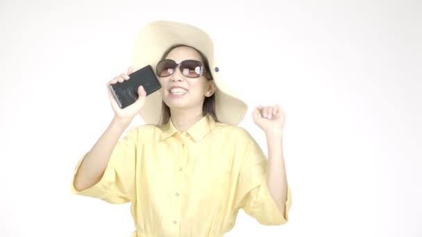 Jonge mooie Aziatische vrouw met zomerhoed zingen met haar telefoon naar camera met witte achtergrond, gelukkig gezicht. Vakantie levensstijl concept.  - Video