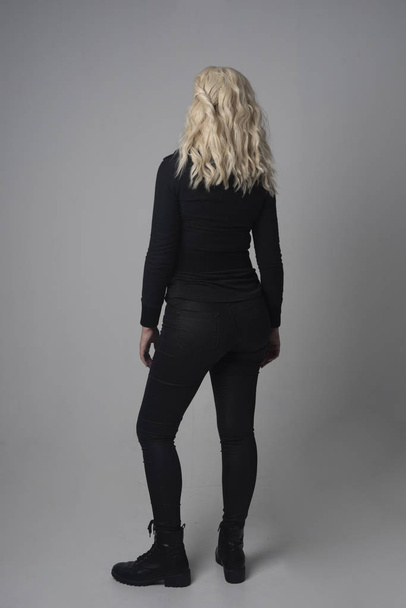volledige lengte portret van een blond meisje dragen moderne zwarte jas en broek, staande pose op de grijze studio achtergrond. - Foto, afbeelding