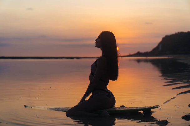 Silhouette sexy girl in swimsuit lying and posing on surf board at beach during sunset - 写真・画像