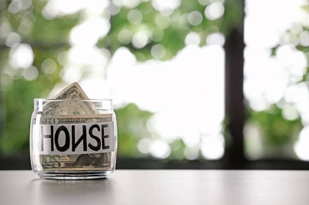 Γυάλινο βάζο με χρήματα και λέξη σπίτι στον πίνακα θολή φόντο, χώρο για το κείμενο - Φωτογραφία, εικόνα