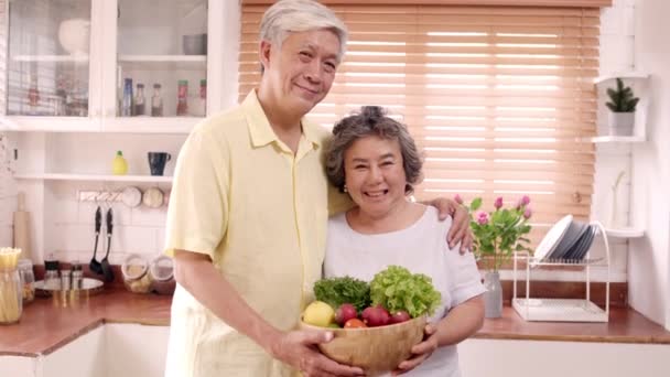 Asian elderly couple feeling happy smiling and holding fruit and looking to camera while relax in kitchen at home. Lifestyle Senior family enjoy time at home concept. Portrait looking at camera. - Video